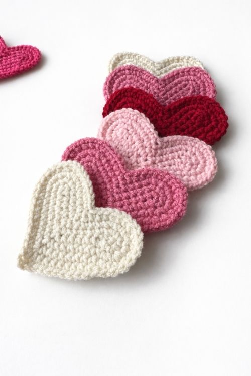 Five crocheted hearts in white, light pink, pink, and red are lined up on a white surface; another pink heart is in the background. Free crochet pattern