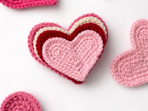 Stack of pink, red, and cream crocheted heart coasters on a white surface; "HOW TO CROCHET A HEART" text is visible. Free crochet pattern