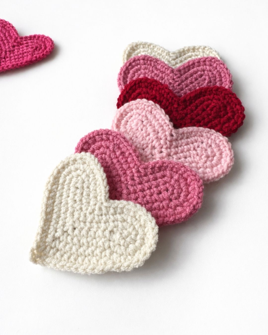 Six crocheted hearts in white, pink, mauve, and red are lined up on white with "HOW TO CROCHET A HEART" text above. Free crochet pattern