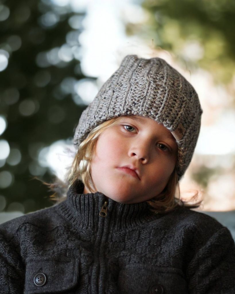 Young child with long blond hair wears a gray ribbed crochet hat and matching sweater, looking down; green blurred background. Free crochet pattern