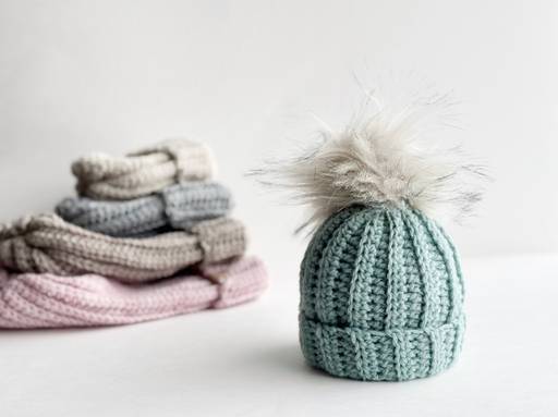 A light blue crochet hat with a fluffy pom-pom is in front, with folded gray and pink items blurred behind on a white surface. Free crochet pattern