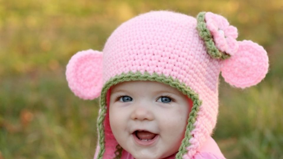 A smiling baby with blue eyes wears an easy, pink crocheted hat with ear flaps, green trim, and a pink flower, outdoors against a blurred grassy background.