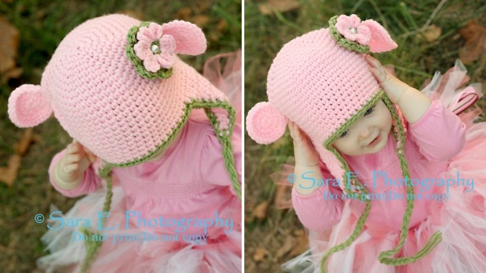 A baby wearing a pink crocheted hat made from worsted weight yarn, featuring green trim, a flower, and small monkey ears, sits outdoors on grass. The cute hat with ear flaps partially covers the babys face.