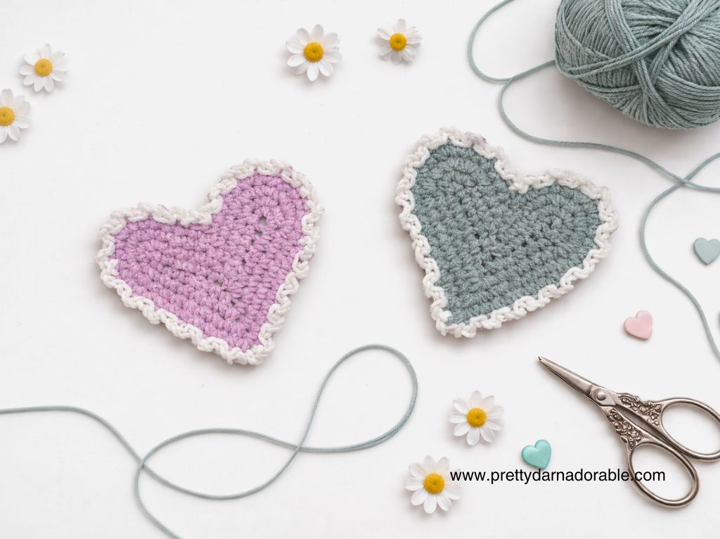 Pink and blue crocheted hearts with white trim sit on a white surface with yarn, flowers, buttons, scissors, and promo text for prettydarnadorable.com. Free crochet pattern
