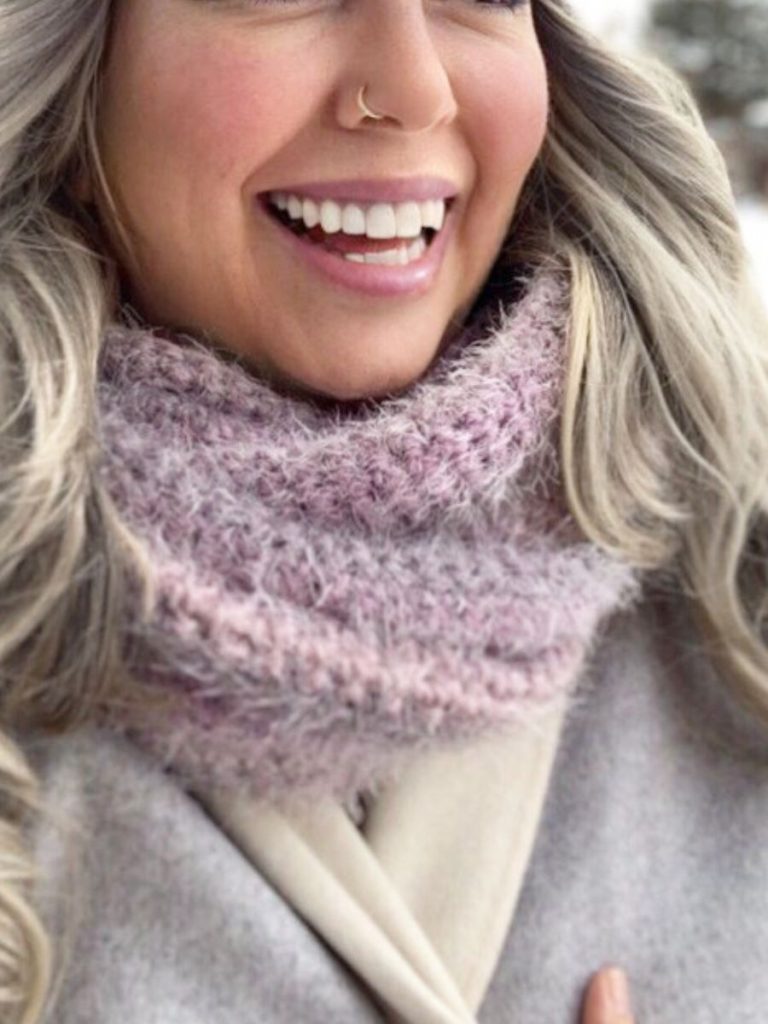 A smiling woman with long, wavy, light-colored hair wears a cozy, textured lavender crochet cowl pattern scarf and a light gray coat. She has a nose ring and is outdoors in a snowy setting.