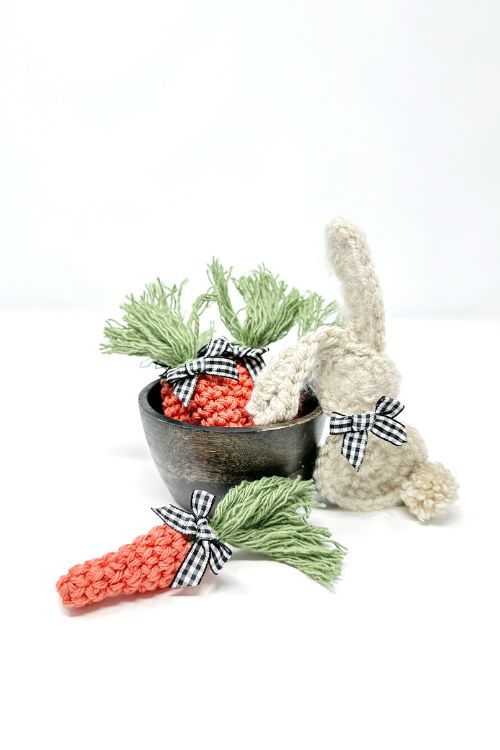 A white crocheted bunny with a checkered bow sits next to a black bowl of crocheted carrots on a white background. Free crochet pattern