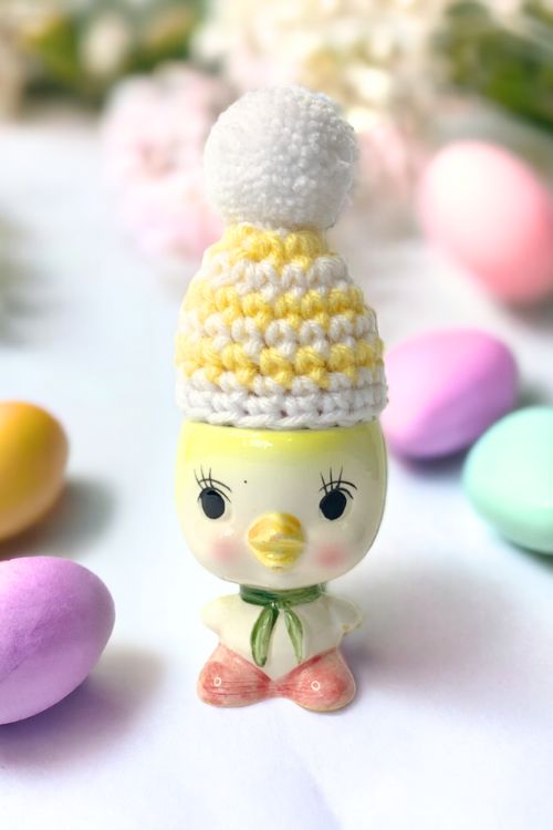 Ceramic chick figurine in a yellow and white yarn hat with pom-pom, surrounded by pastel eggs, next to Free Crochet Carrot Pattern text. Free crochet pattern