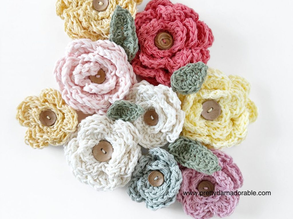 A collection of crocheted flowers in pastel shades of pink, yellow, cream, and white with wooden button centers and green leaves, arranged in a cluster&mdash;perfect accents for projects like a free crochet bucket hat pattern.