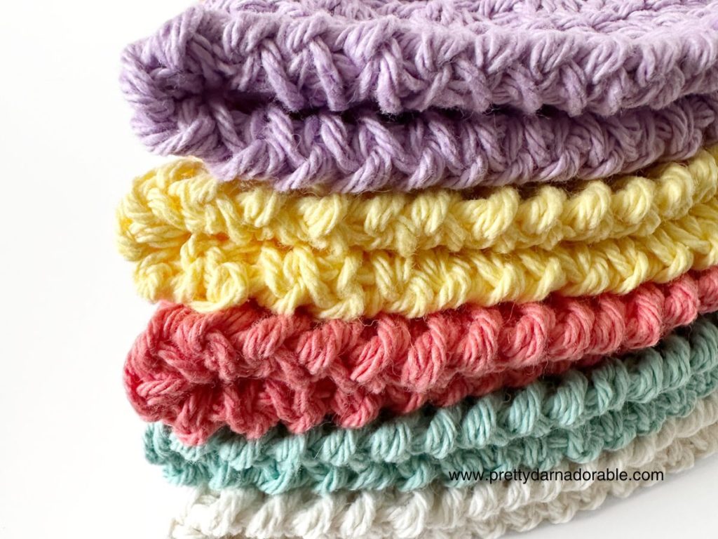 A neatly stacked pile of crocheted blankets in pastel colors, including purple, yellow, pink, mint green, and white, displaying textured stitches on a white background&mdash;perfect inspiration for your next free crochet bucket hat pattern.