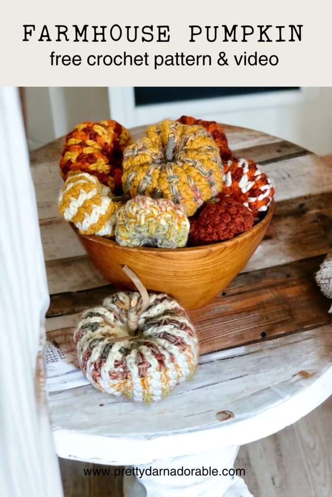 A wooden bowl filled with colorful, chunky crocheted pumpkins sits on a rustic wooden table, with one multicolored crocheted pumpkin in front. Text above offers a free crochet pattern and video for an easy crochet pumpkin—perfect for fall crochet decor. Free crochet pattern