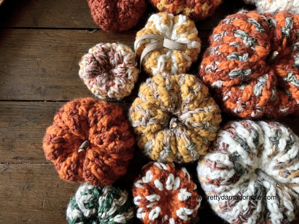 A collection of handmade, chunky knit and crochet pumpkins in various autumn colors—orange, yellow, white, and multicolor—arranged on a rustic wooden surface. Perfect for adding cozy fall crochet charm to your decor. Free crochet pattern