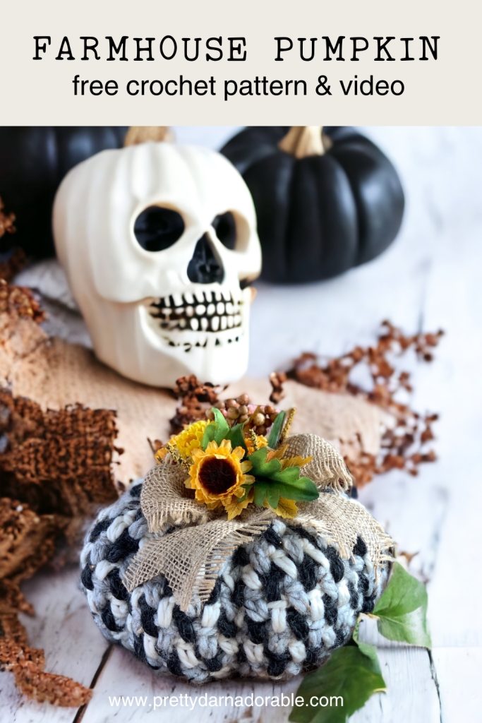 A black and white crochet pumpkin with a burlap bow and faux sunflower sits before a white skull and black pumpkins on a rustic surface. Text reads: Farmhouse Pumpkin, easy crochet pumpkin free pattern & video. Free crochet pattern