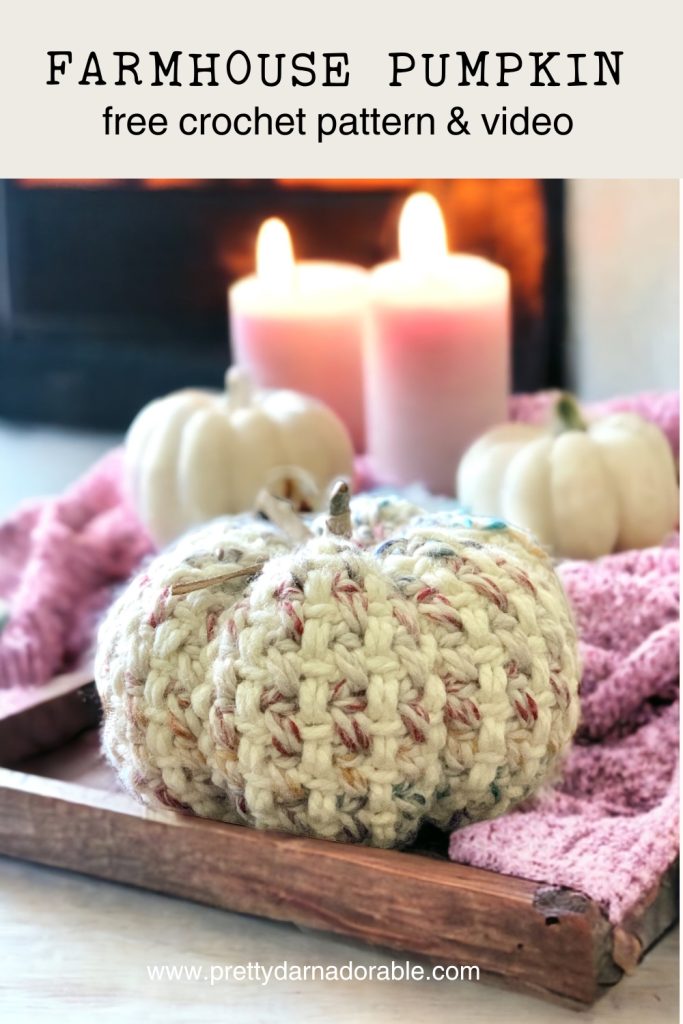 A crochet pumpkin sits on a wooden tray with a pink knitted cloth, surrounded by white pumpkins and lit candles. Text reads: “FARMHOUSE PUMPKIN free crochet pattern & video”—a cozy fall crochet idea!. Free crochet pattern