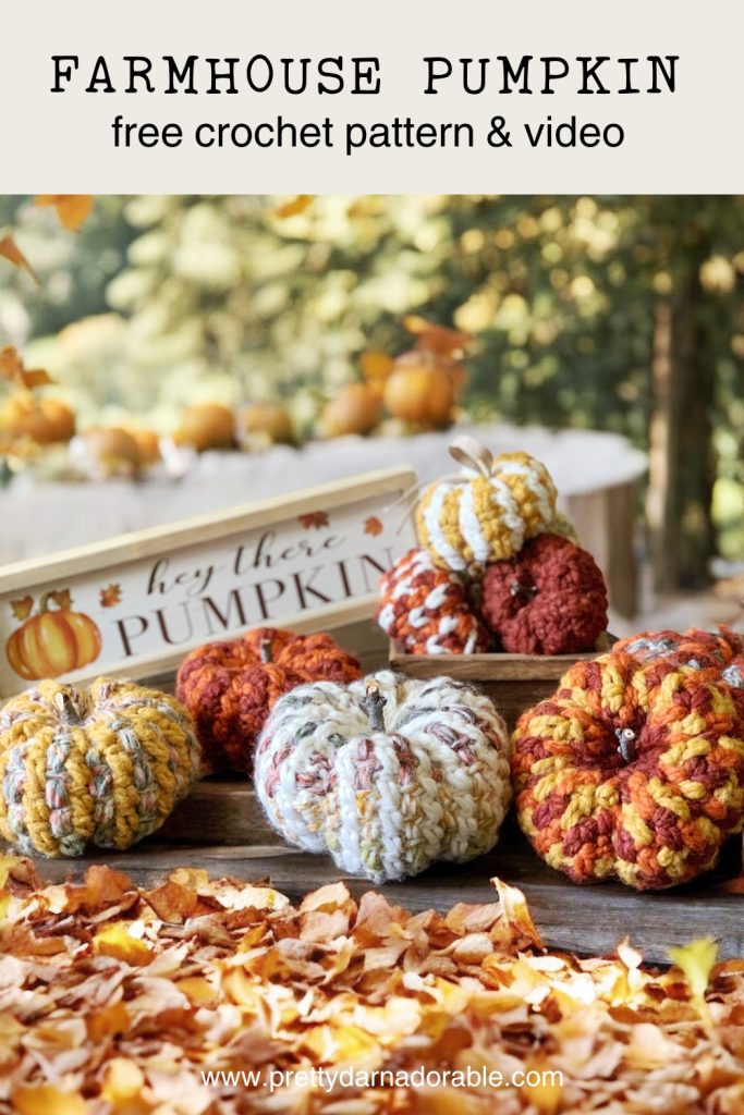 Several easy crochet pumpkins in autumn colors are displayed among fallen leaves, with more pumpkins and a wooden box labeled hey there PUMPKIN in the background. Trees with fall foliage are visible behind the display. Free crochet pattern