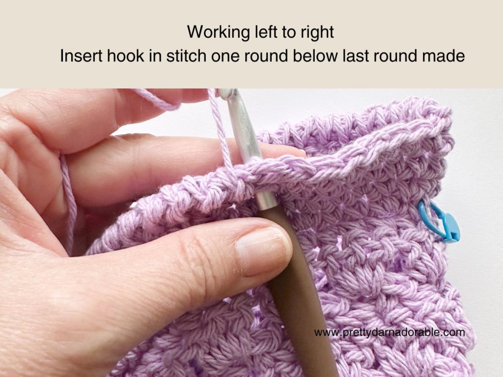 A hand holding a crochet hook, inserting it into a purple crocheted piece. Instructions above read: Working left to right. Insert hook in stitch one round below last round made, ideal for following a free crochet bucket hat pattern.