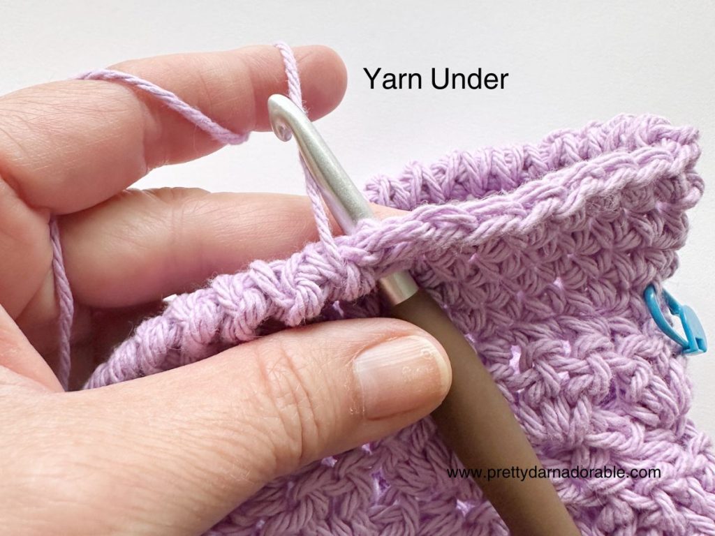 A close-up of a hand holding a crochet hook, inserting it into lilac yarn stitches, with the text Yarn Under above and www.prettydarnadordable.com on the bottom right, featuring a free crochet bucket hat pattern.