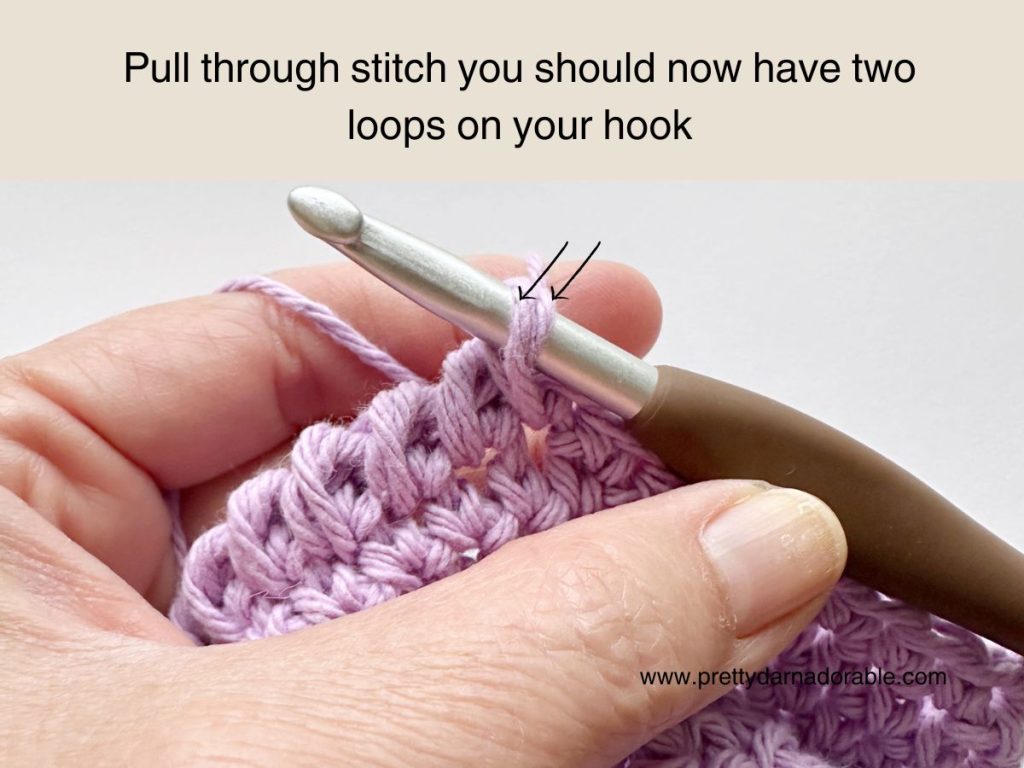 A close-up of a hand holding a crochet hook with two loops of purple yarn on it. Text above reads, Pull through stitch&mdash;you should now have two loops on your hook in this free crochet bucket hat pattern.