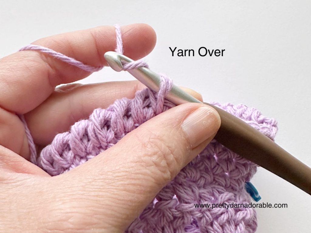 A close-up of a hand holding a crochet hook and purple yarn, showing the yarn over step in crochet. Perfect for beginners following a free crochet bucket hat pattern. The words Yarn Over are labeled near the hook.