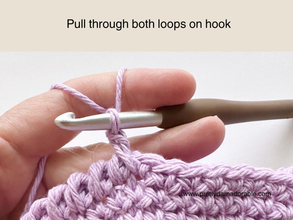 A hand holding a crochet hook with light purple yarn, following the instruction Pull through both loops on hook above a partially crocheted fabric from a free crochet bucket hat pattern.