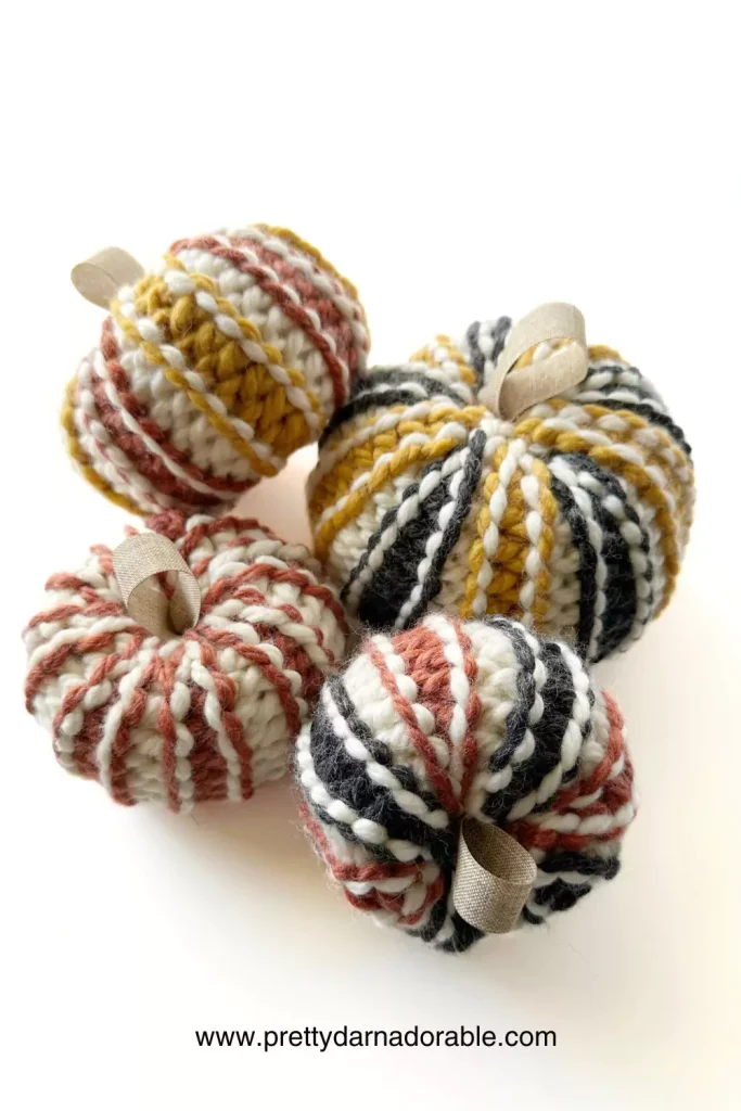 Four small, chunky knit pumpkins in cream, yellow, black, and rust stripes on a white surface; free pattern at prettydarnadorable.com. Free crochet pattern
