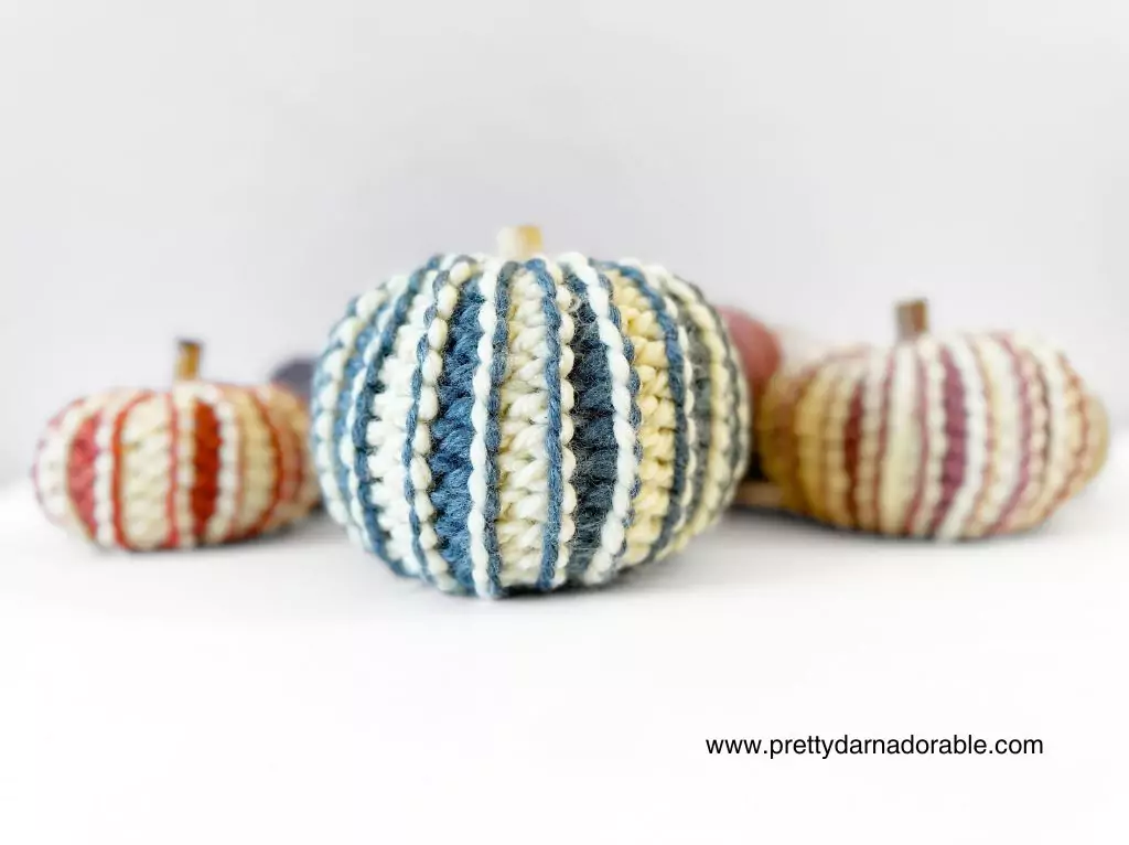 Four small pumpkins in multicolored crochet yarn sit on white; front has blue, cream, yellow stripes. Website promo text in corner. Free crochet pattern