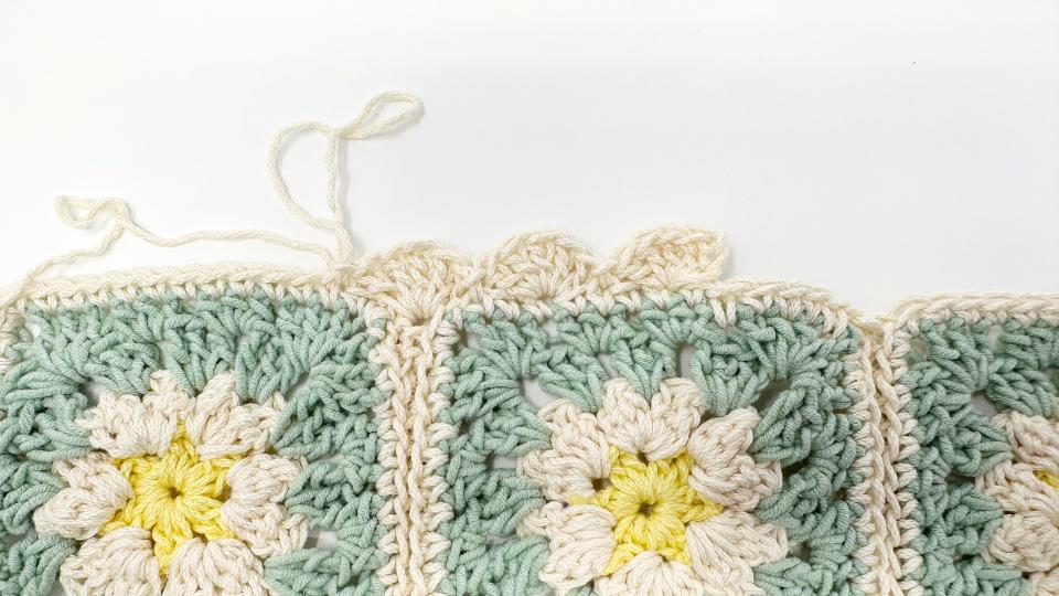 Close-up of crocheted granny squares with a flower pattern in pastel yellow, cream, and green yarn—perfect for kitchen decor. The unfinished cream-colored edge and loose yarn ends are shown on a white background. Free crochet pattern available.