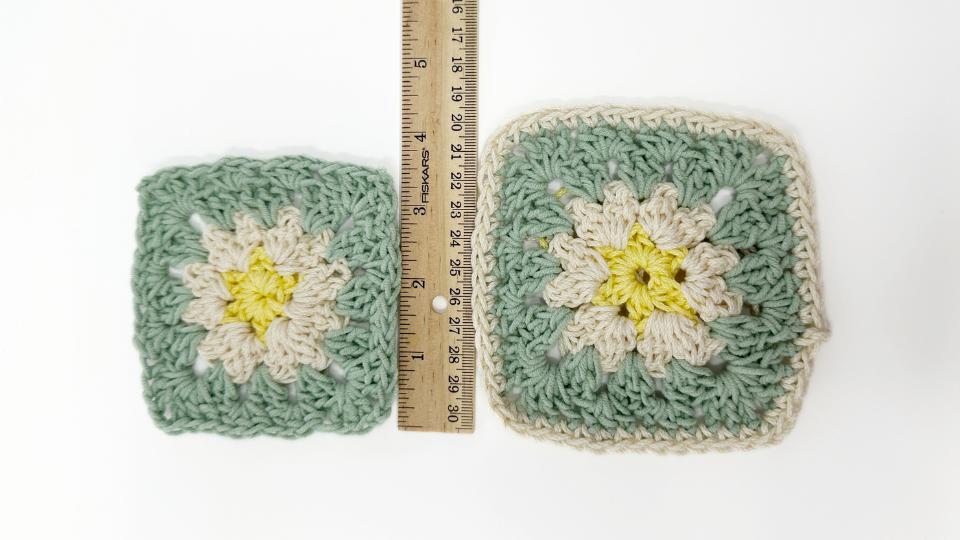 Two crocheted granny squares in green, cream, and yellow are placed next to a wooden ruler on a white background. One square is slightly smaller than the other, showing a size comparison—perfect for illustrating crochet placement in projects.