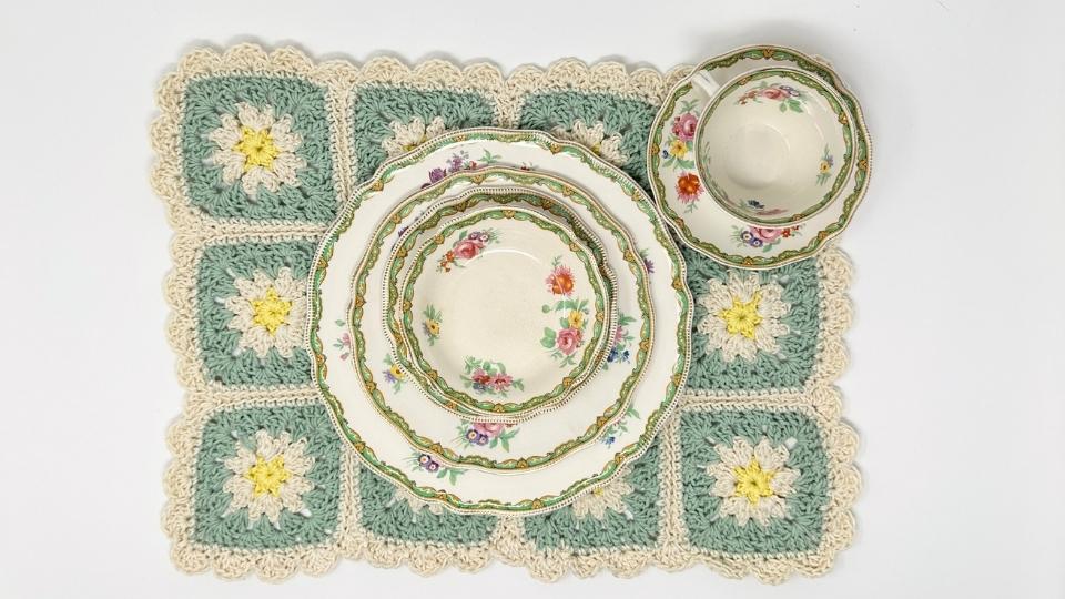 A floral-patterned tea set with plates and a teacup sits on a green and cream granny square placemat made of white and yellow flower designs, perfect for those seeking granny square placemat free patterns.