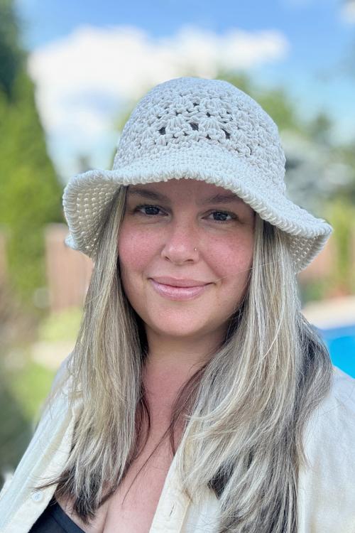 A woman with long blonde hair smiles at the camera while wearing a light crocheted sun hat, made from a free crochet pattern, and a cream-colored shirt. The background shows greenery, a fence, and a blurred swimming pool.