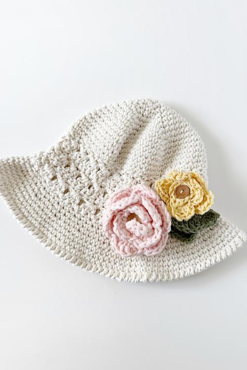 A cream-colored crocheted hat decorated with a pink and a yellow crocheted flower with green leaves, displayed on a white background. Includes free crochet pattern and video tutorial.