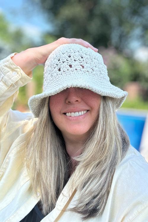 A smiling woman with long blonde hair wears a crocheted white hat that partially covers her eyes. Perfect for ladies and baby, this look shines outdoors against blurred greenery and blue sky&mdash;great inspiration for your next video tutorial.
