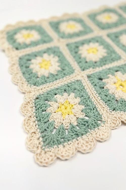 A close-up of a crocheted blanket featuring flower motifs with yellow centers, cream petals, green backgrounds, and a scalloped cream border. The joined square sections highlight thoughtful crochet placement, perfect for cozy kitchen decor.