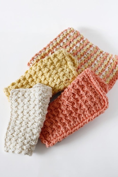 A stack of three crocheted dishcloths on a white background. The top one is white, middle is pale yellow, and bottom is pink, all with a textured pattern.