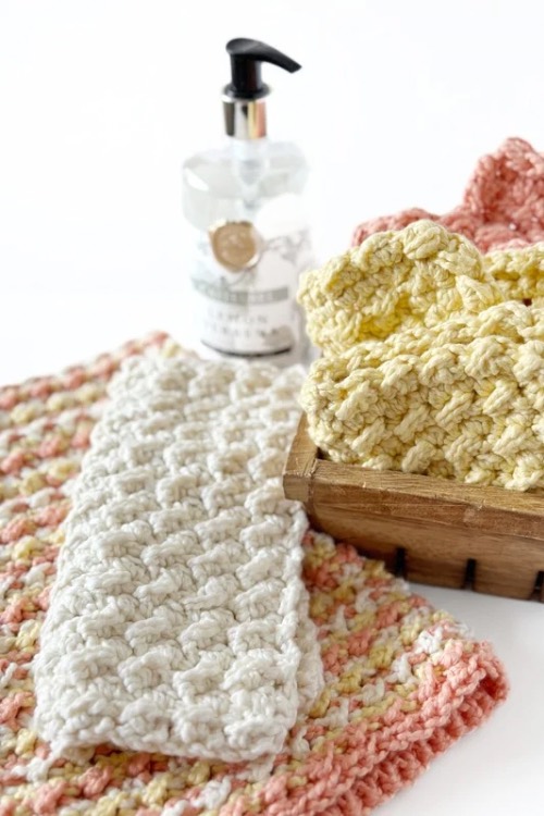 Textured crochet cloths in soft white, pink, and yellow tones are arranged with a wooden box and a bottle in the background, conveying warmth and coziness.