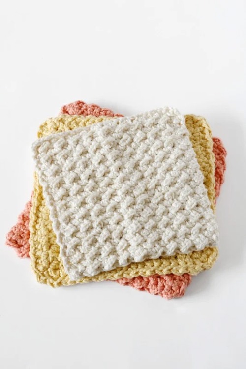 A stack of three crocheted dishcloths on a white background. The top one is white, middle is pale yellow, and bottom is pink, all with a textured pattern.