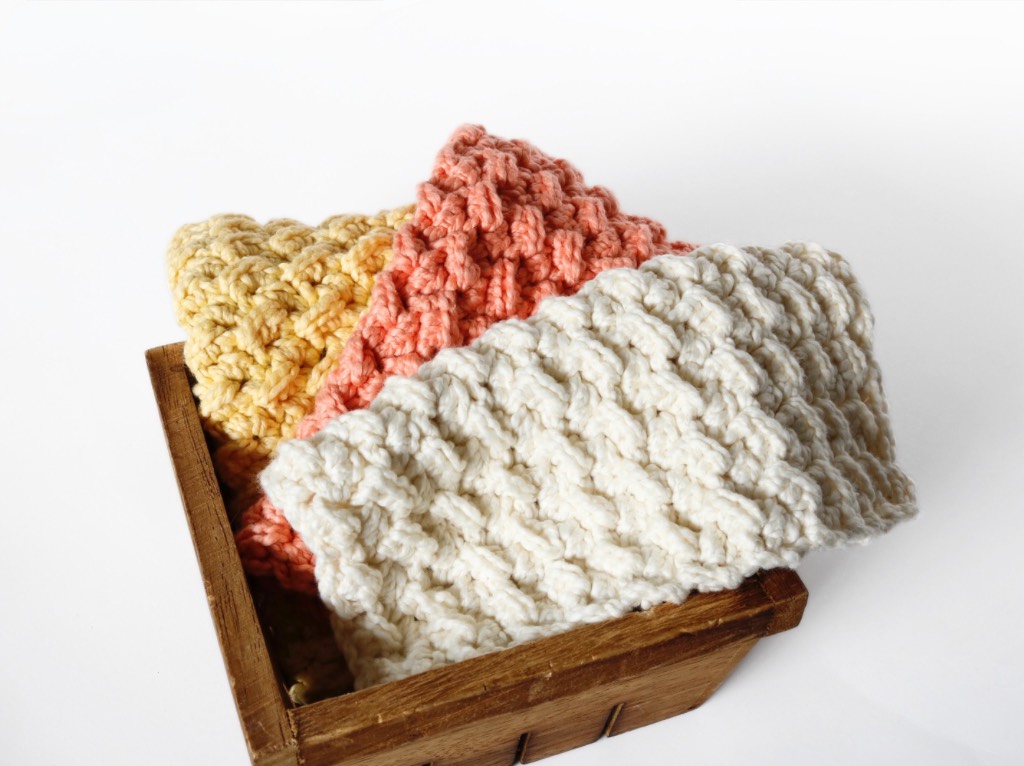 Textured crochet cloths in soft white, pink, and yellow tones are arranged with a wooden box and a bottle in the background, conveying warmth and coziness.
