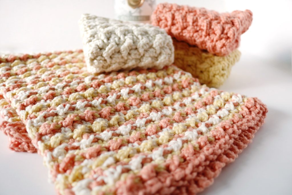 Soft, crocheted washcloths in pastel shades of white, peach, and yellow are layered against a bright background, conveying warmth and coziness.