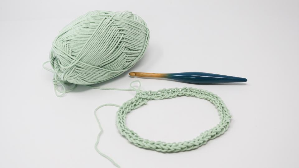 A ball of light green yarn, a crochet hook, and a circular row of stitches are arranged on a white surface—perfect for starting a simple granny stitch crochet beanie.