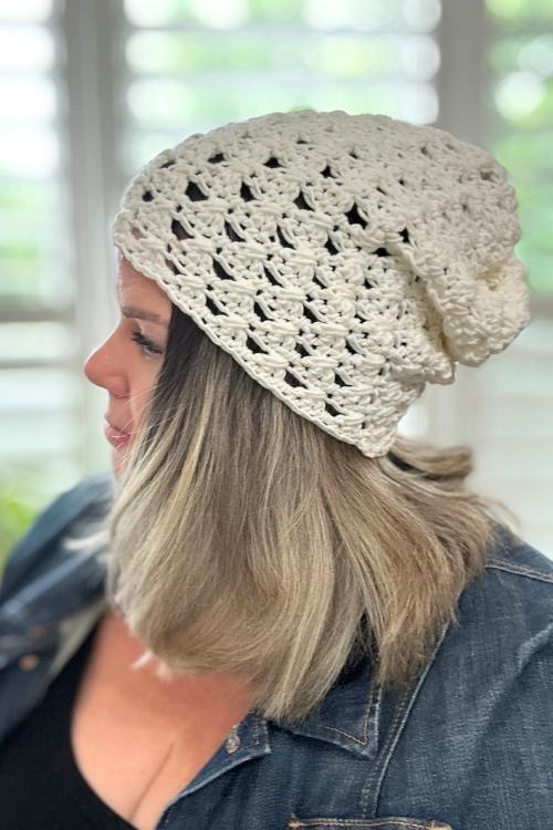 A woman with shoulder-length, light brown hair wears a loose, white crochet granny stitch beanie and a denim jacket. She is facing left, with a softly blurred background featuring window blinds.