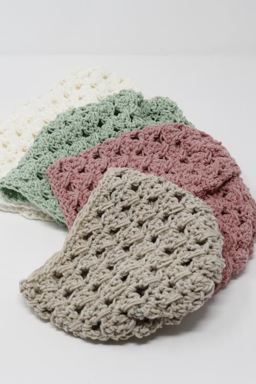 Four crocheted washcloths in neutral colors—cream, green, pink, and beige—are layered on a white surface. Their chunky, textured stitch pattern echoes the classic look of a crochet granny stitch beanie.