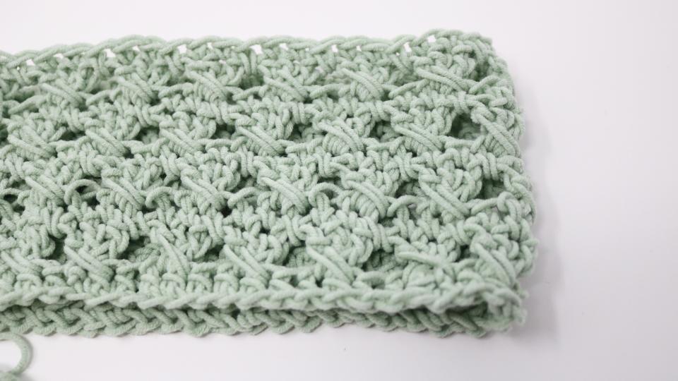 A close-up of a light green crocheted fabric with an open, textured pattern using the crochet granny stitch, displayed on a white surface. The thick, soft yarn creates a delicate, lacy appearance perfect for a cozy beanie or hat.
