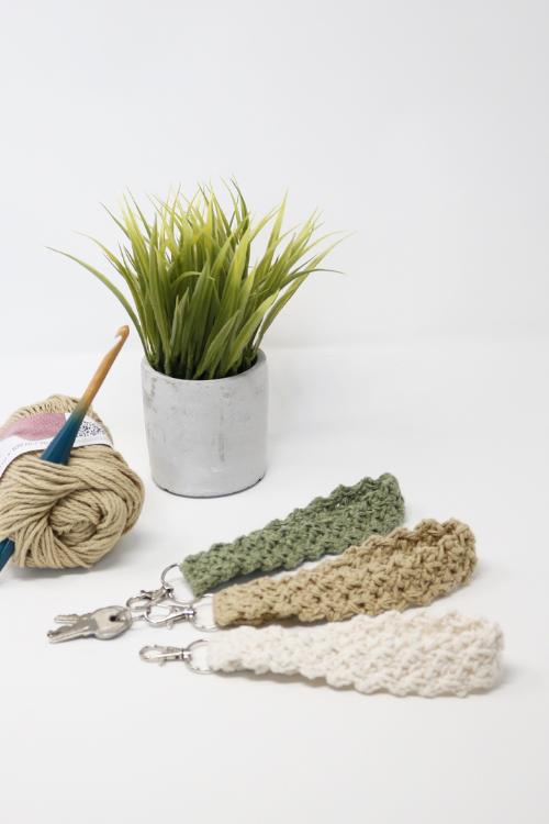 A potted green plant sits beside a crochet hook, a ball of beige yarn, and three keychains made using free crochet keychain patterns in green, tan, and cream colors on a white surface.