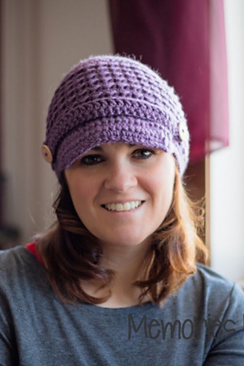 A woman with brown hair smiles while wearing a purple crocheted hat with a button on the side. She is dressed in a gray top and is indoors with a blurred background.