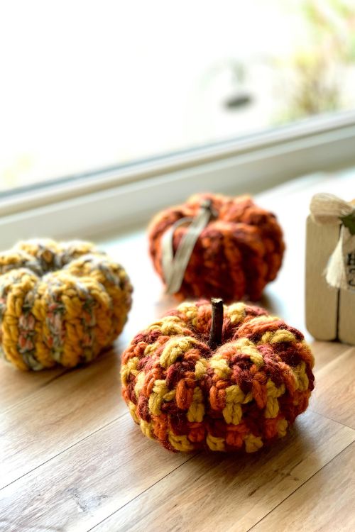 Three crocheted pumpkins in autumn colors—orange, yellow, and multicolored—sit on a wooden surface near a window, creating a cozy fall decoration. Free crochet pattern