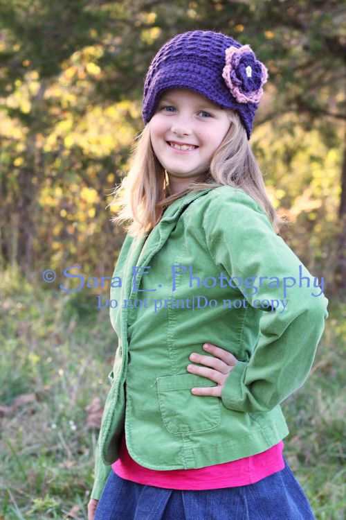 A young girl smiles outdoors, wearing a green jacket, pink shirt, blue denim skirt, and a purple knit hat with a flower. She stands in profile with one hand on her hip, with trees and sunlight in the background.
