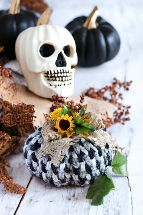 A white skull and two black pumpkins are displayed on a rustic wooden surface. In front, a chunky pumpkin with a striking black-and-white pumpkin pattern is decorated with burlap, sunflowers, and greenery for a festive Halloween arrangement. Free crochet pattern