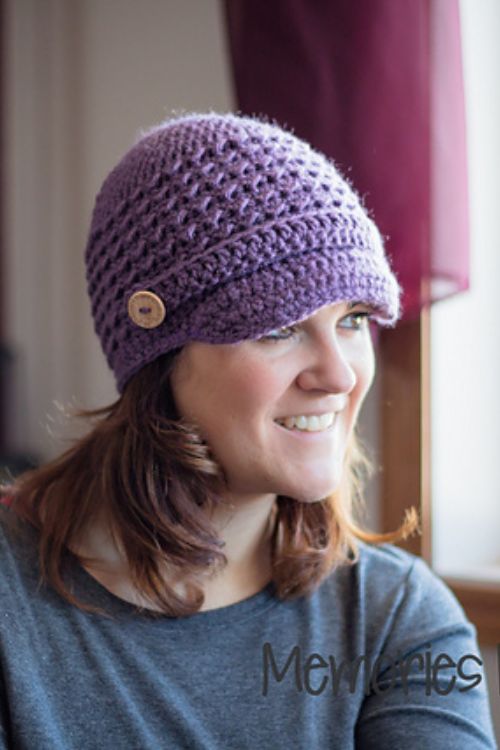 A woman wearing a purple crocheted hat with a wooden button on the side smiles and looks to her right. She is dressed in a grey top and is indoors, with maroon curtains in the background.