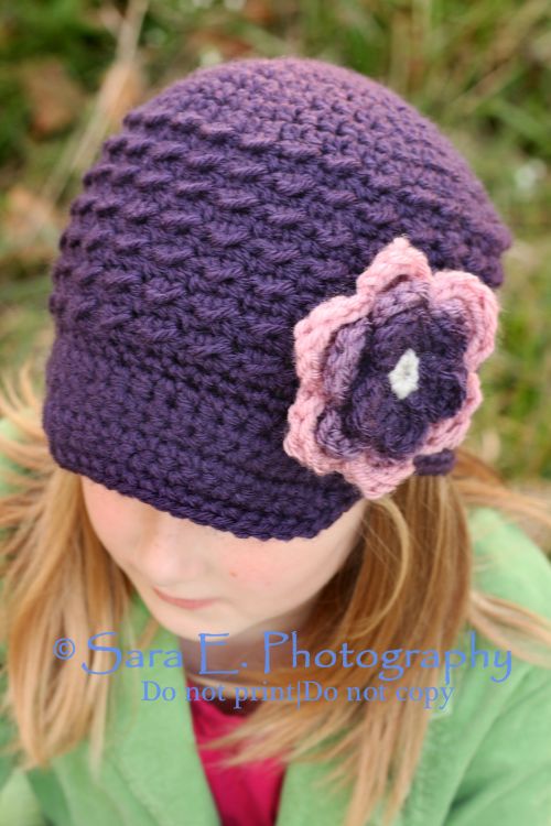 A girl wearing a purple crocheted hat with a pink and purple flower on the side, seen from above. She has light brown hair and is dressed in a green jacket.