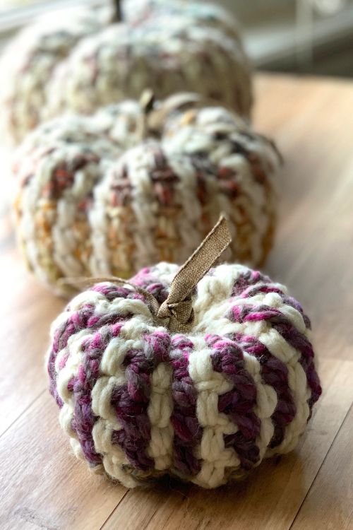 Three crochet pumpkins—cream, purple, and brown—sit on wood with chunky yarn texture and fabric stems. No visible text. Free crochet pattern