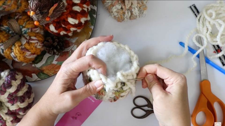 Person sewing thick white yarn with a needle; scissors, ruler, and colorful knits rest on a green leaf-patterned plate.